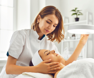 cosmetologist directing lamp to woman for facial treatment