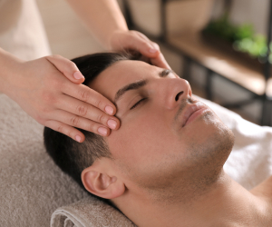 Person receiving a head massage in a spa setting, lying on a towel-covered surface with hands gently massaging their scalp.