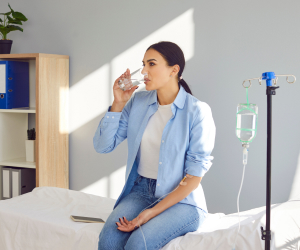 A person sitting on a medical bed, receiving IV therapy, and drinking water. The room has a bookshelf and a potted plant.