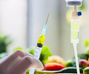 A gloved hand holds a syringe filled with yellow liquid, positioned near an IV drip. Blurred vegetables are visible in the background.