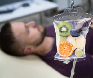 A person lying down with an IV bag containing various fruits, including kiwi, orange, grape, and peach, suggesting a concept of vitamin infusion therapy.