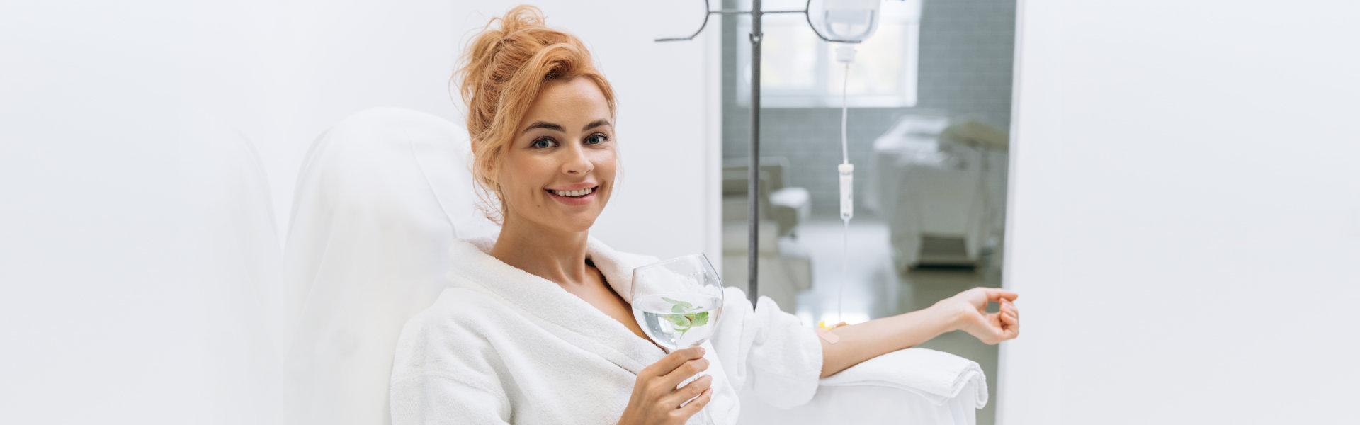 Person in a white robe relaxing in a spa-like setting, holding a glass of water with mint, receiving an IV drip.