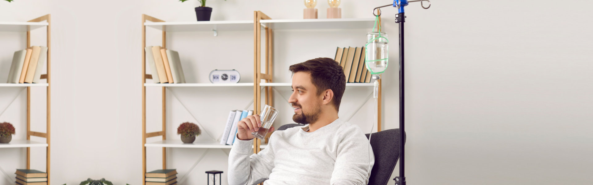 A person sitting in a chair receiving an IV drip, holding a glass of water, with a bookshelf and plants in the background.