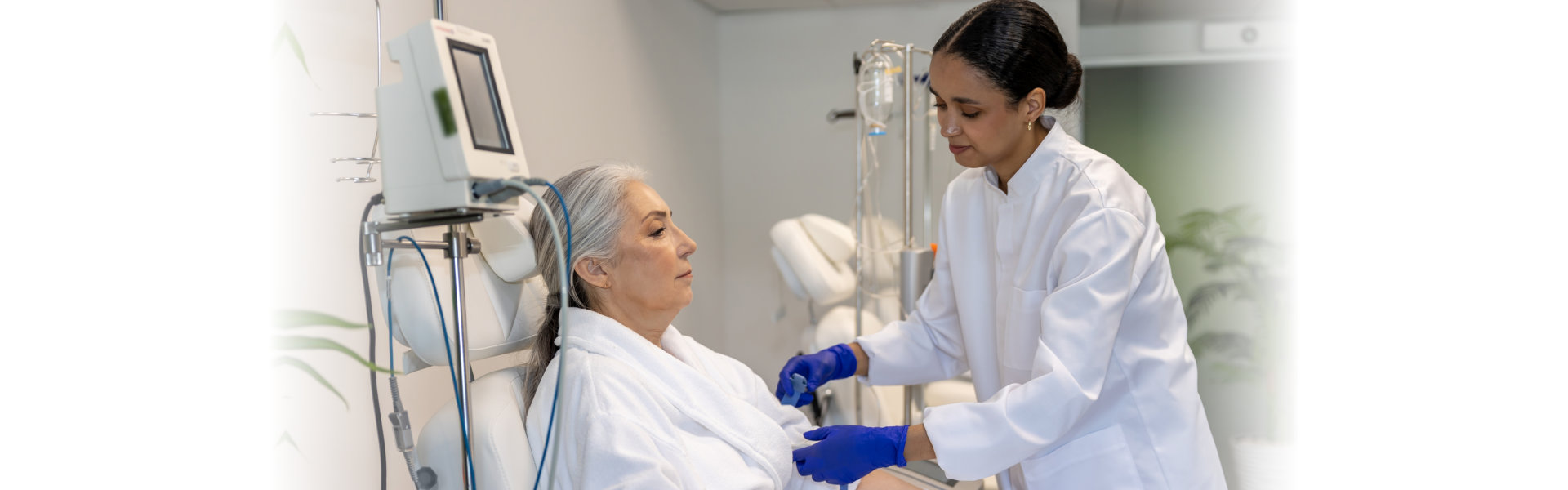 A healthcare professional in a white coat and blue gloves administers an IV drip to a patient seated in a medical chair.