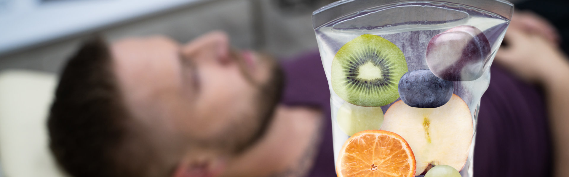 A person lying down with an IV bag containing various fruits, including kiwi, orange, grape, and peach, suggesting a concept of vitamin infusion therapy.