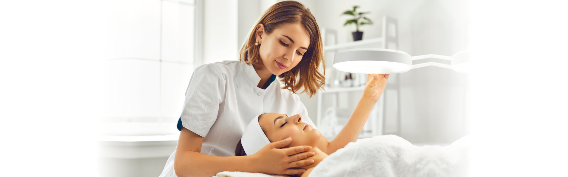 cosmetologist directing lamp to woman for facial treatment