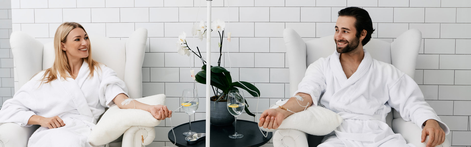 Two people in white bathrobes sit in armchairs receiving IV therapy, with a table holding drinks and a plant between them.